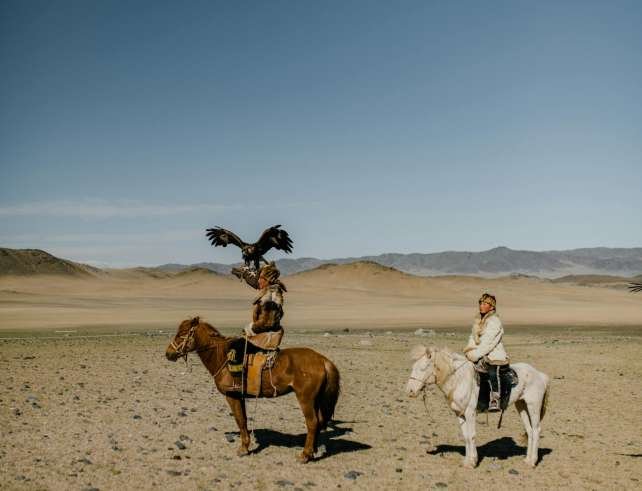 Side view of ethnic people in traditional clothes riding horses in dry sandy desert covered with pebbles