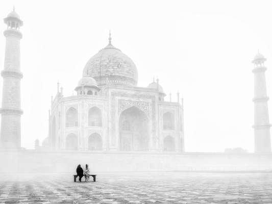 Two people sit before the foggy Taj Mahal, capturing its serene beauty.