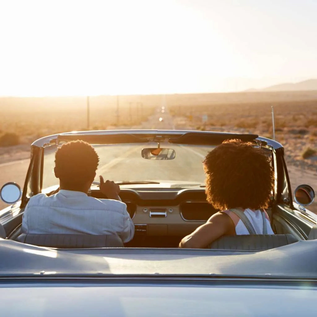 Rear View Of Couple On Road Trip Driving Classic Convertible Car Towards Sunset